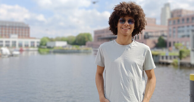 Jhorman stands smiling in front of a lake, behind there are buildings